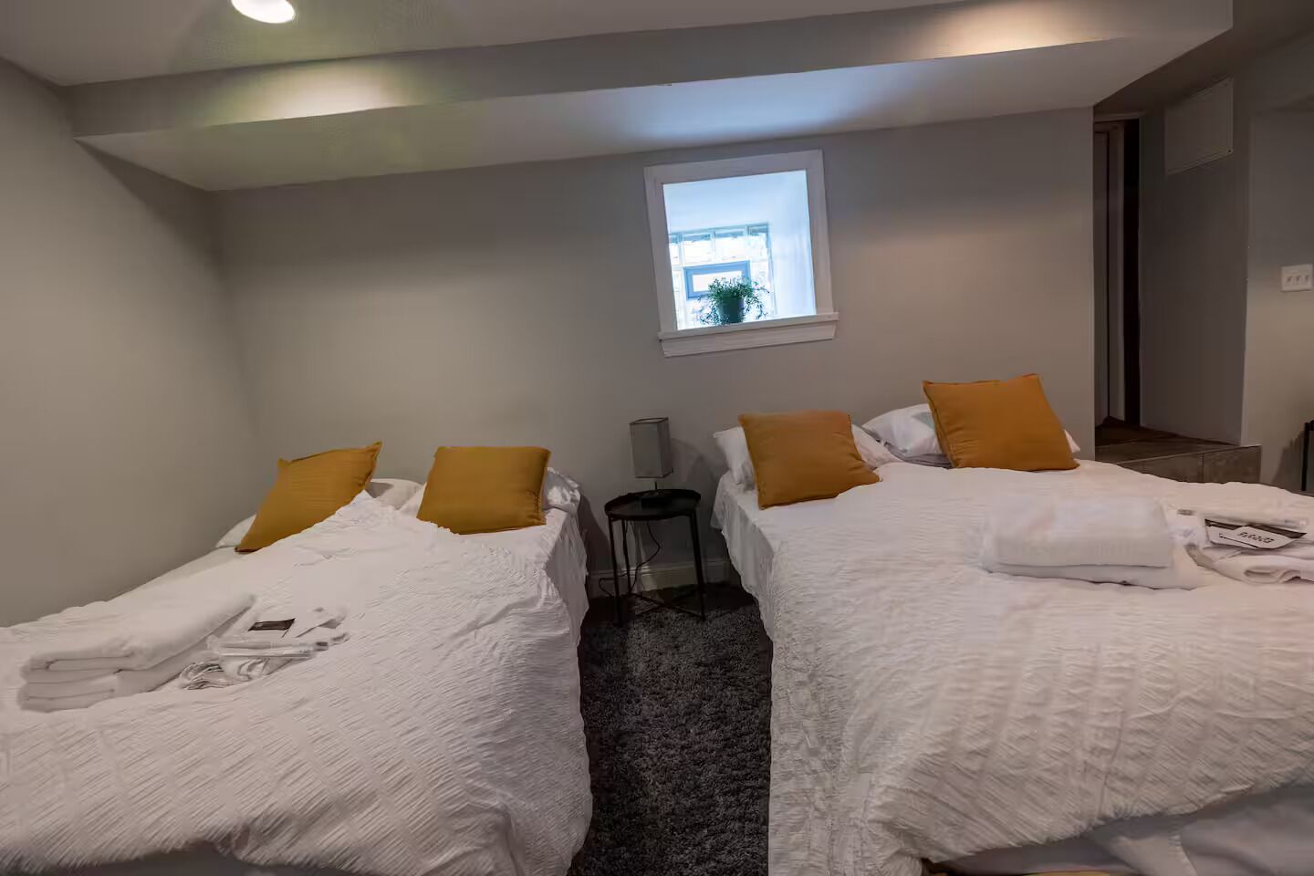 Hotellihuone, jossa on kaksi sänkyä, molemmissa valkoiset vuodevaatteet ja sinapinväriset tyynyt, pieni pöytä ja ikkuna, josta tulee luonnonvaloa.