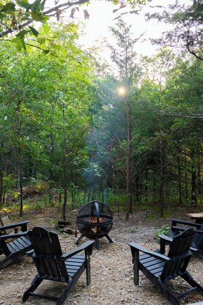 A forested area with a fire pit and Adirondack chairs.
