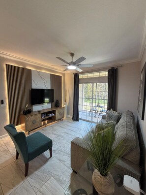 A modern living room with a flat-screen TV, a ceiling fan, and a large window with curtains. - Modern/beautiful Mediterranean Oasis Near Disney (Kissimmee)