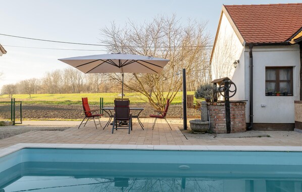 Un área de piscina con una mesa y sillas bajo una sombrilla, un muro de ladrillo y un edificio con tejado de tejas.