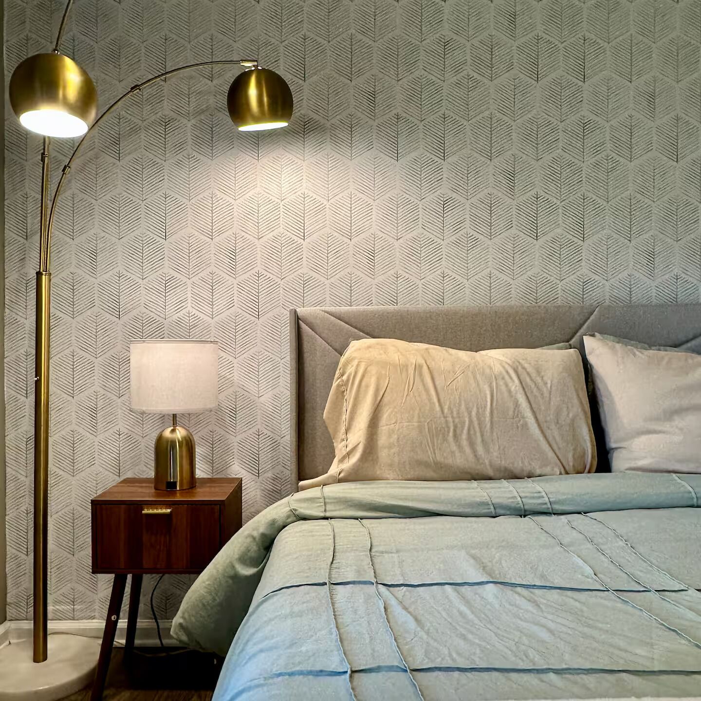 Un dormitorio con un papel tapiz estampado, una cama con colcha azul y una lámpara de pie con pantalla dorada.