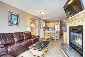 A living room with a leather sofa, a coffee table, a fireplace, and a kitchen in the background. - Cozy 2BR Stay w/ Balcony | Walk to Lifts + Hot Tub (Keystone)