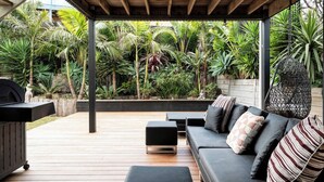 A covered outdoor seating area with a wooden deck, a black sofa with patterned cushions, and a hanging chair. - Sweeping Ocean Views (Torquay)