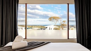 A hotel room with a large window offering a view of a coastal landscape, a neatly made bed, and a rolled towel on the bed. - Sweeping Ocean Views (Torquay)