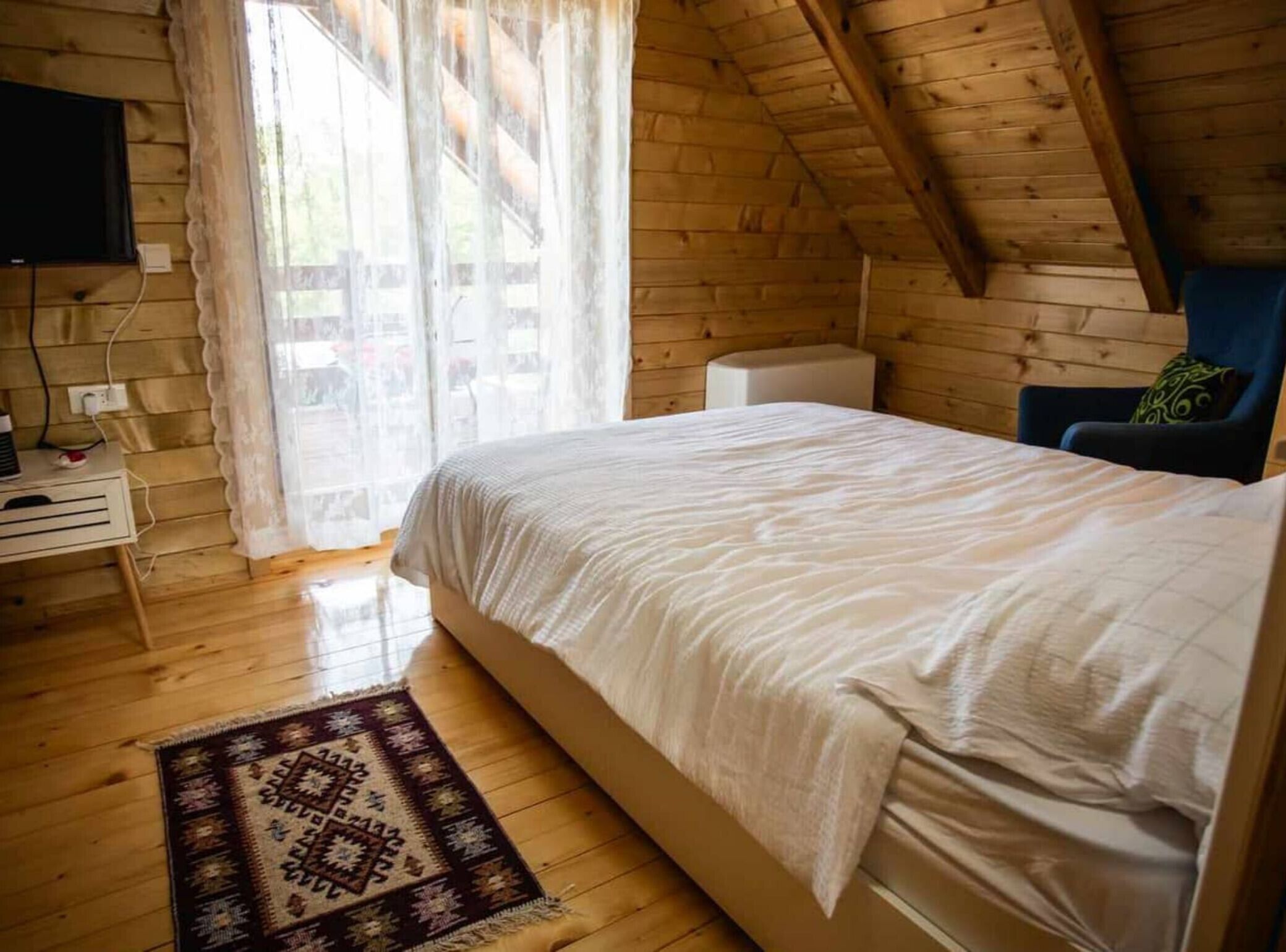 Una habitación acogedora con techo de madera, una cama con colcha blanca, un pequeño escritorio con computadora y una alfombra estampada.