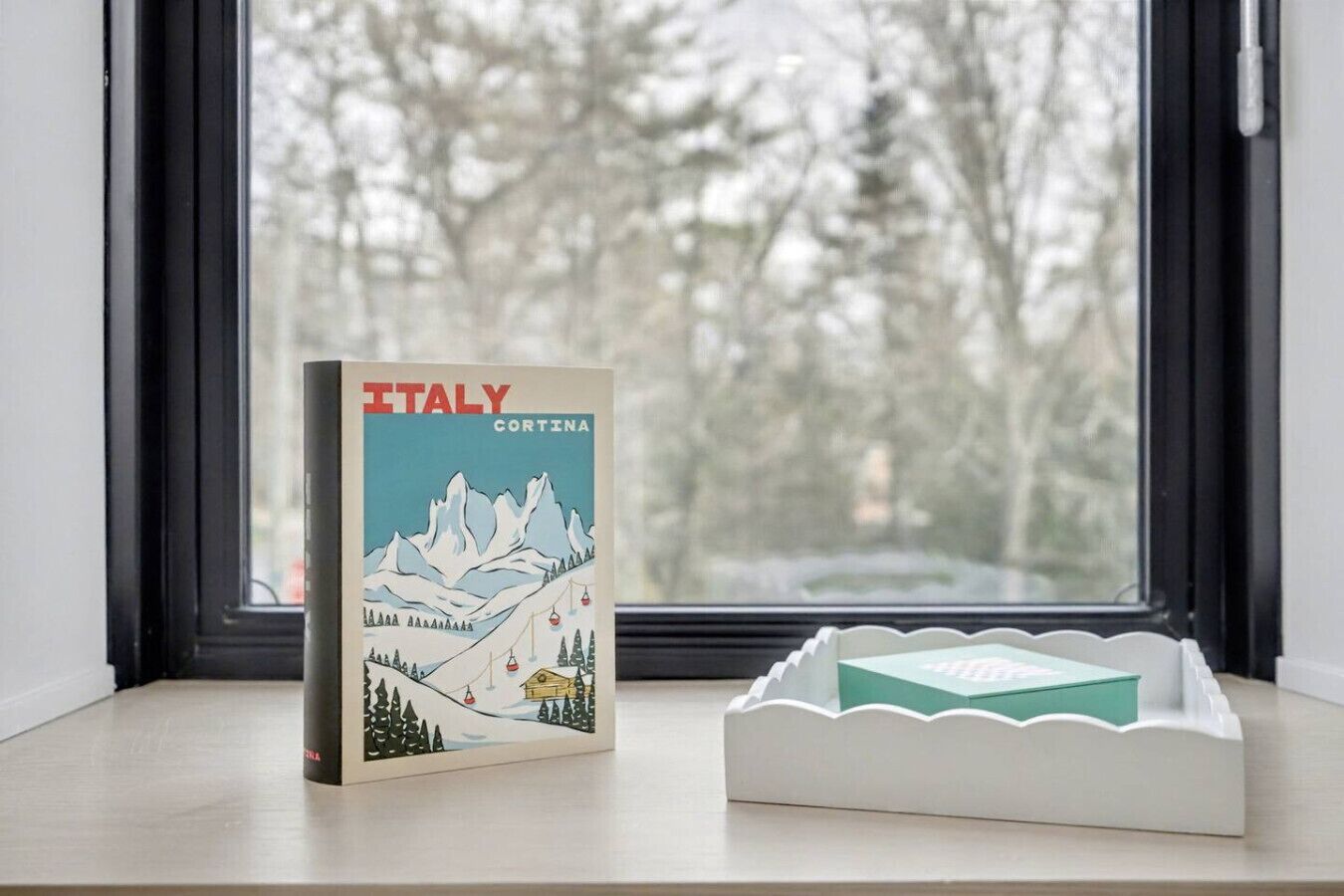 Un libro con el título 'ITALY CORTINA' sobre un alféizar con vista a árboles.