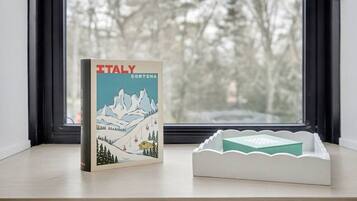 Un libro con el título 'ITALY CORTINA' sobre un alféizar con vista a árboles.