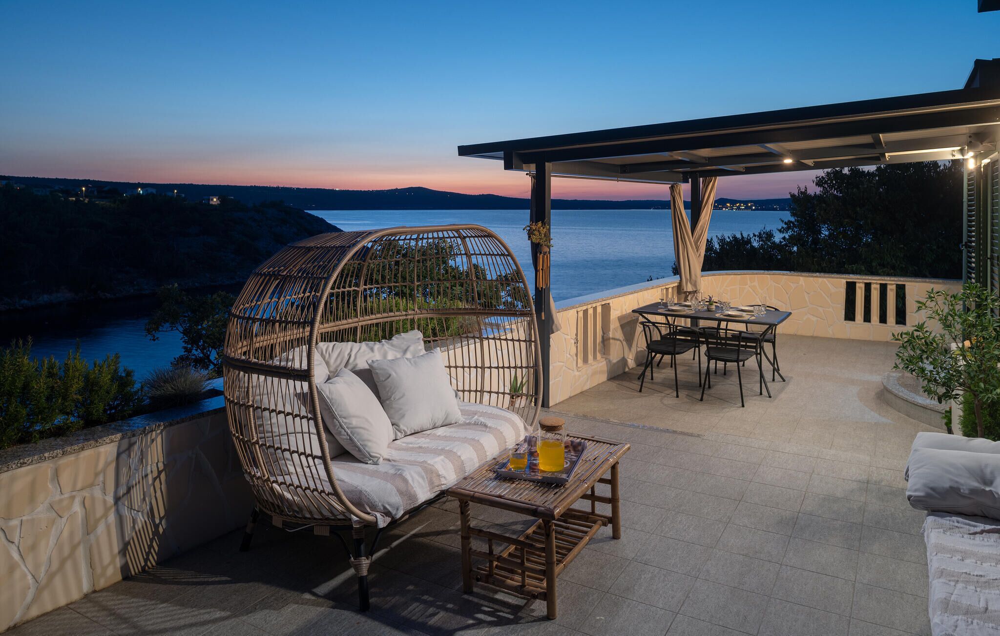 A terrace with a wicker sofa, a dining table, and chairs, overlooking a body of water at dusk.