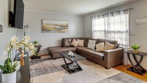 A living room with a brown leather sofa, a black coffee table, and a TV mounted on the wall. There are potted plants and a painting on the walls. - Near Beaches, Base & Bay! Hampton Home w/ Backyard (Hampton)