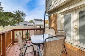 Une terrasse couverte avec une table en verre et des chaises en osier, une terrasse en bois et une vue sur les maisons voisines.
