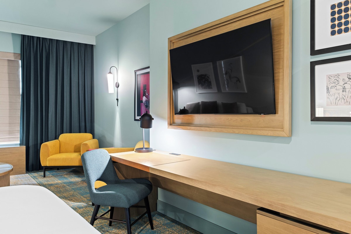 Una habitación de hotel con un escritorio de madera, un sillón amarillo, una silla azul, una cama, un ventanal con cortinas y obras de arte en las paredes.