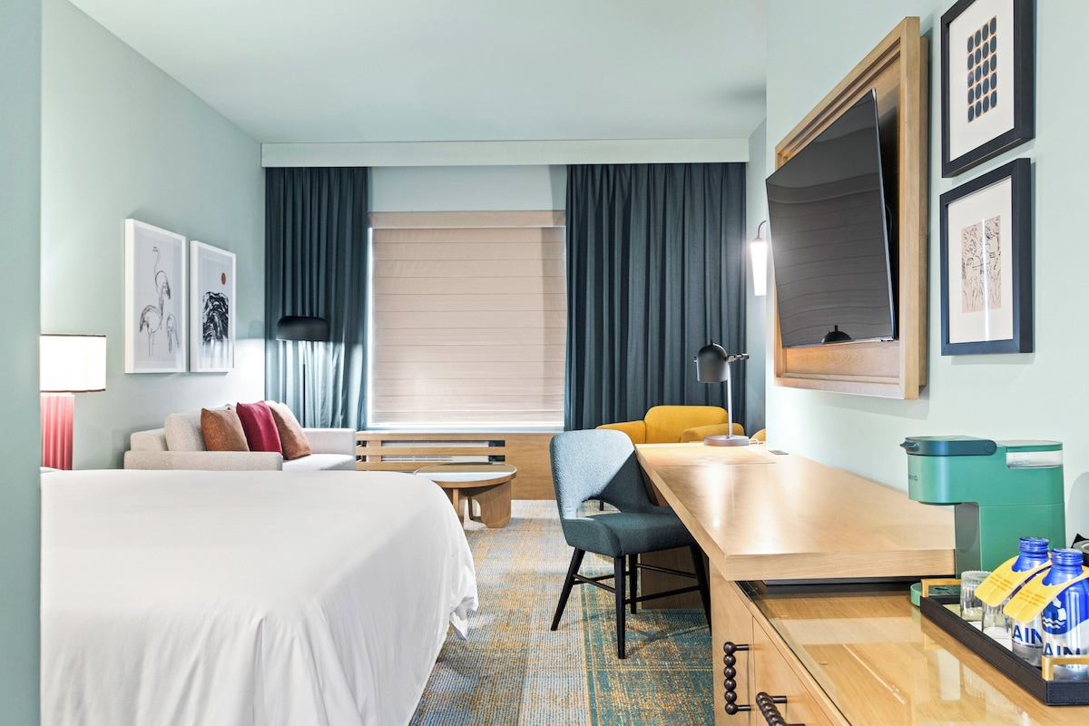 Habitación de hotel con cama, escritorio, silla, mesita, banco, sofá y ventana con cortinas.