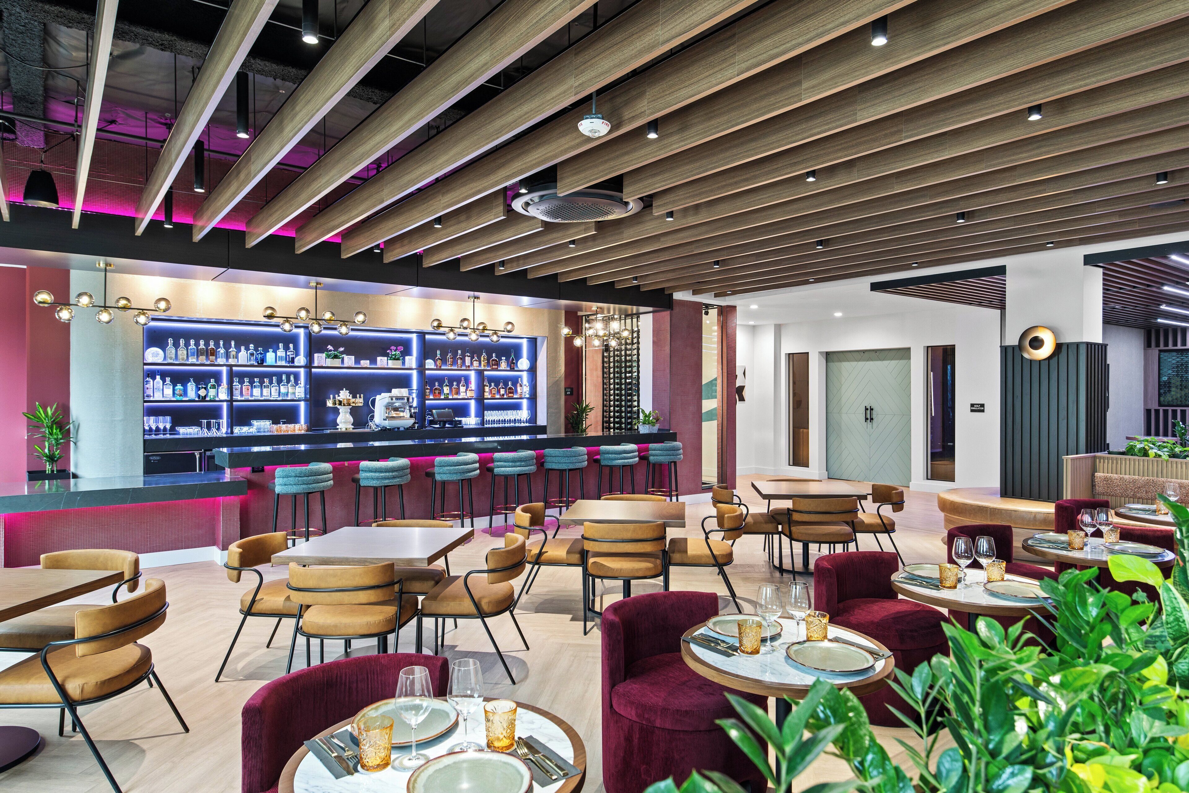 A modern restaurant with wooden ceiling beams, bar area with shelves of bottles, and seating arrangements including round tables and cushioned chairs.
