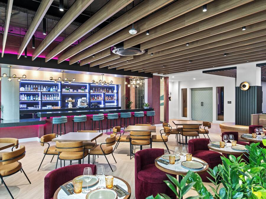 A modern restaurant with wooden ceiling beams, bar area with shelves of bottles, and seating arrangements including round tables and cushioned chairs.