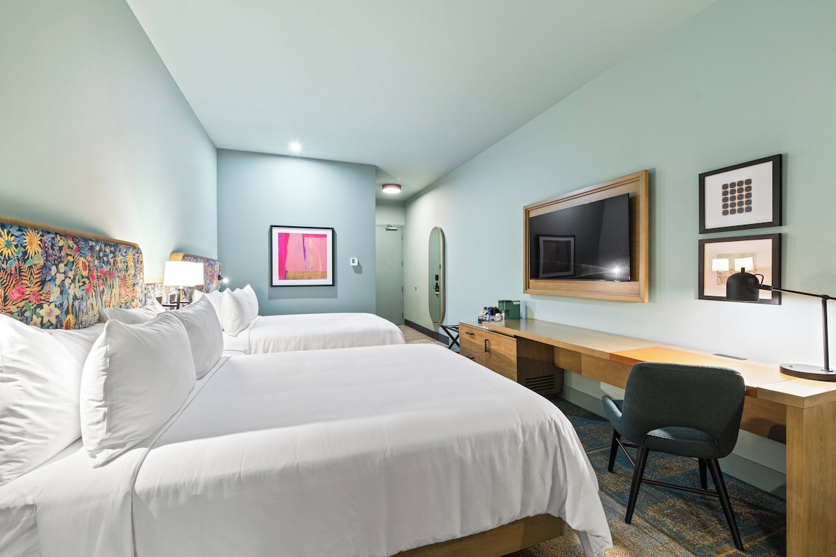 Habitación de hotel con dos camas, un escritorio, una silla, un televisor y obras de arte en las paredes.