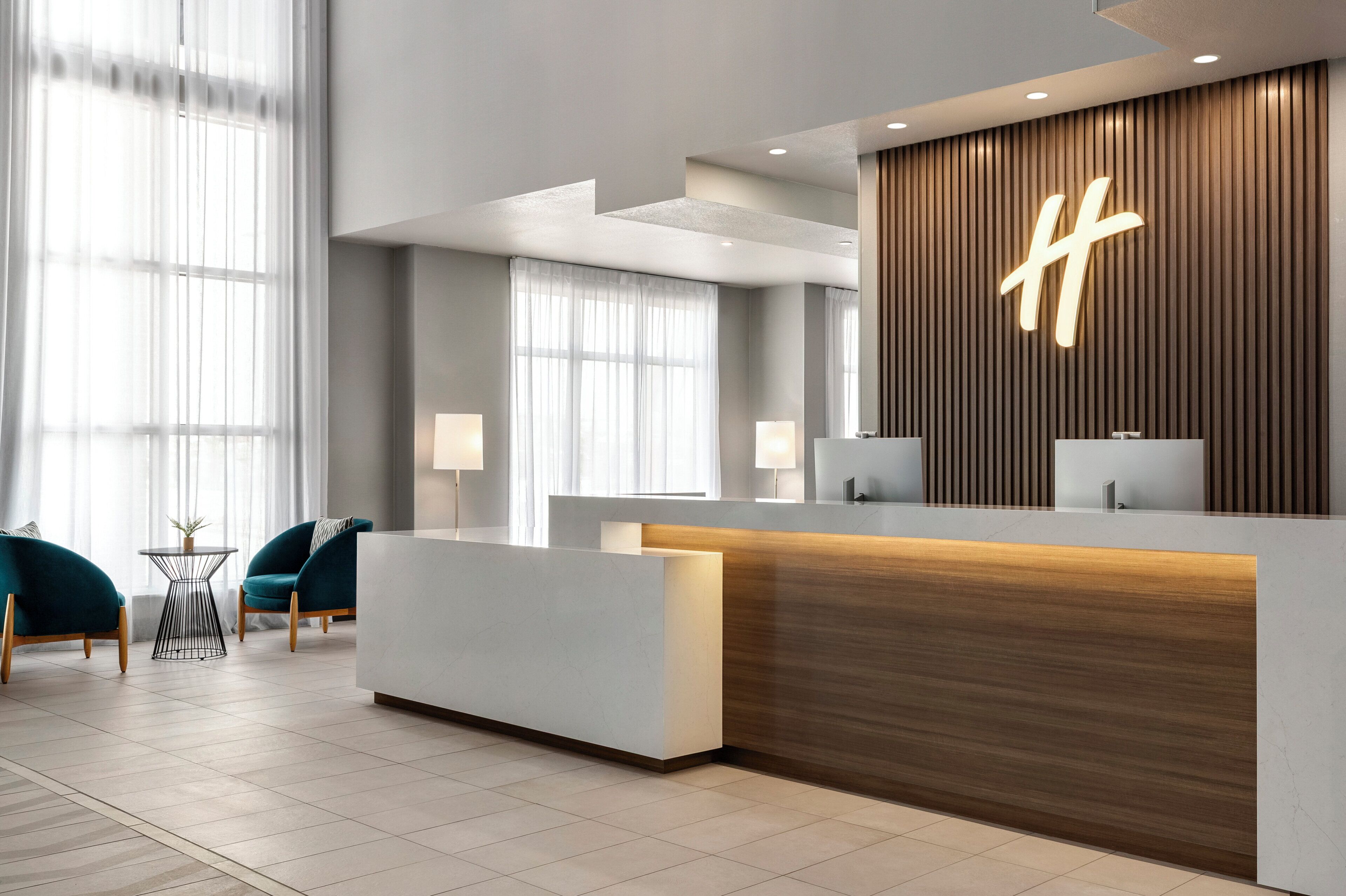 A modern reception area with a wooden accent wall, a large illuminated 'H' sign, and a seating area with a teal chair and a small table.