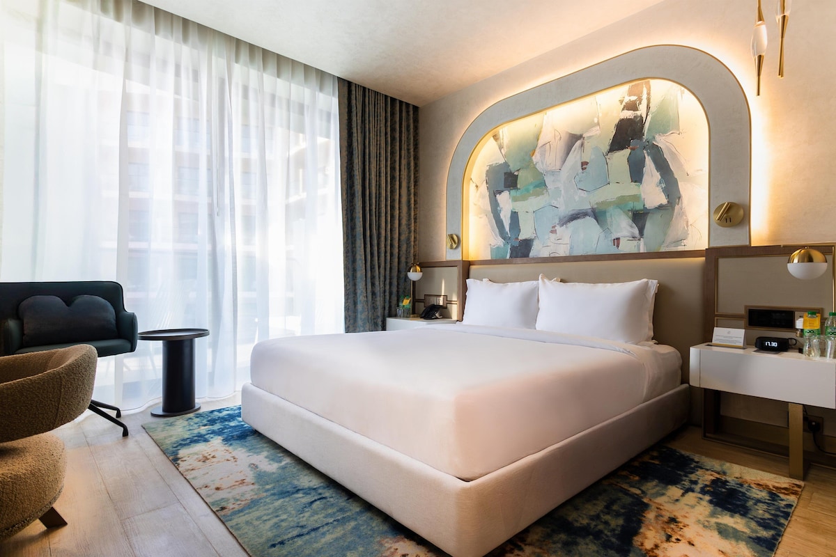 Ein modernes Hotelzimmer mit einem großen Bett, einer Sitzecke mit einem Sessel, einem Nachttisch und einem großen, abstrakten Gemälde über dem Bett.