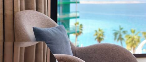 A chair with a cushion, a window with curtains, and a view of the ocean.