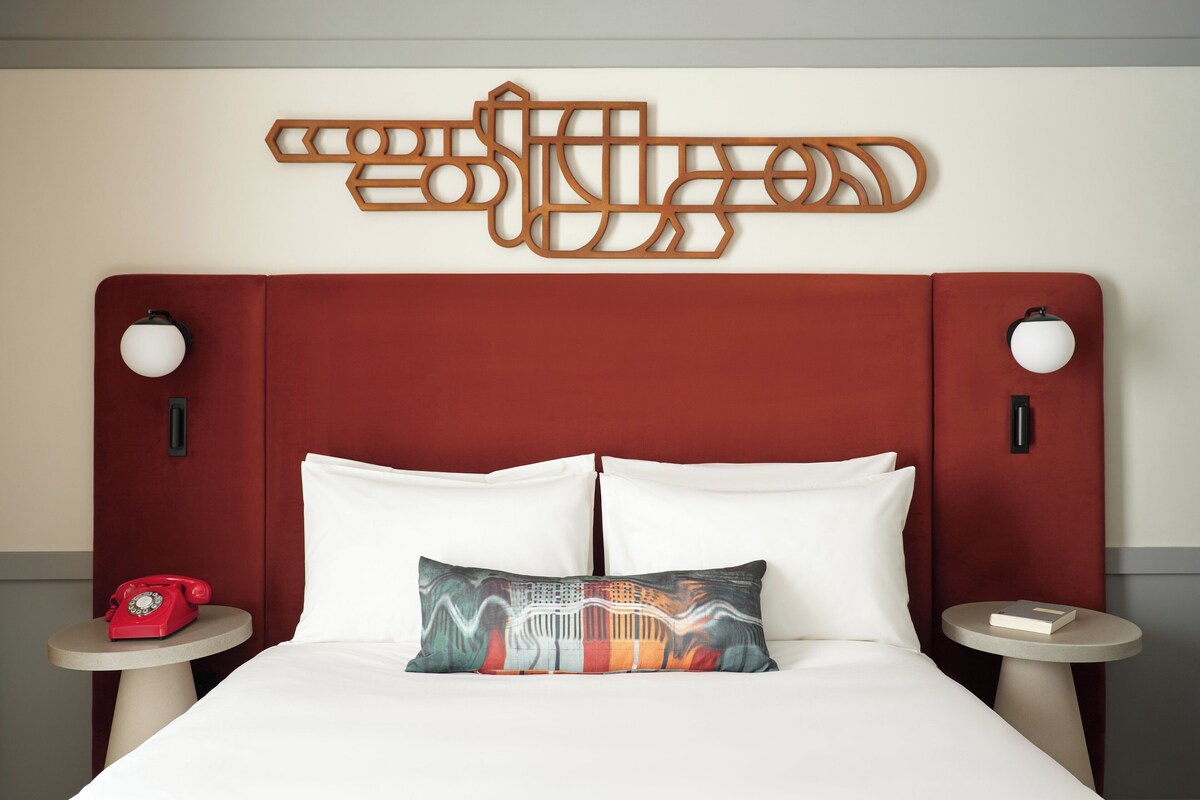 Una habitación de hotel con cabecera roja, ropa de cama blanca, un cojín decorativo, un teléfono rotativo rojo y una obra de arte abstracto colgada en la pared.