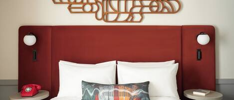 A hotel room with a red headboard, white bedding, a decorative pillow, a red rotary phone, and a wall-mounted abstract art piece.