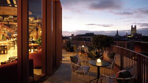 Un espacio al aire libre con mesas y sillas, vista a un paisaje urbano y un edificio con una torre.