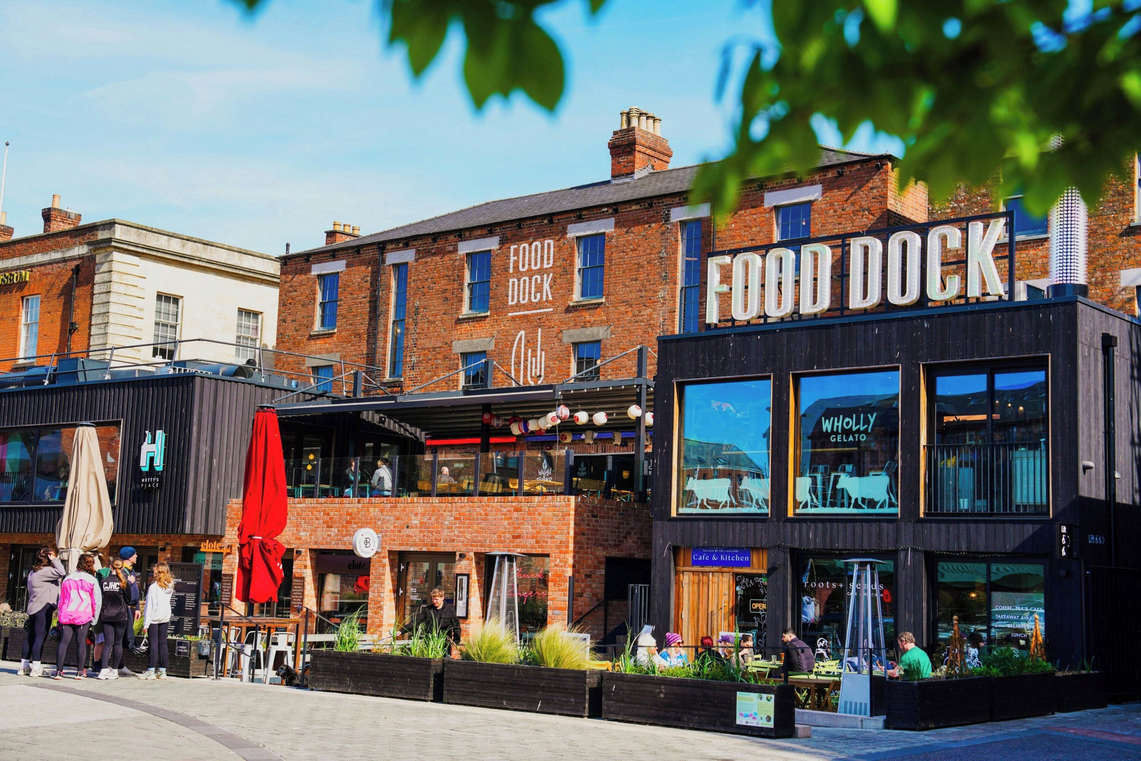 Una calle con una hilera de edificios, incluyendo un restaurante llamado 'Food Dock' y otro con un letrero que dice 'Café & Kitchen'.