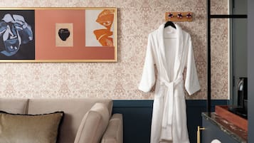 Una stanza con carta da parati decorata, un quadro incorniciato, un accappatoio bianco su un appendiabiti, un divano beige con un cuscino a righe e un mobile scuro.