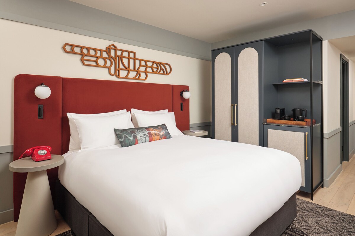A modern hotel room with a large bed, a red telephone, a side table, and a wardrobe with a mirror.
