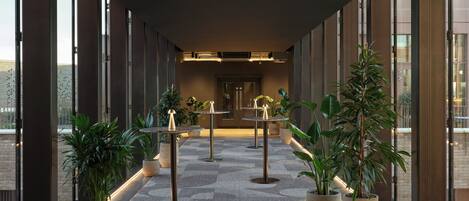 A corridor with potted plants, tables, and a view of the city through large windows.
