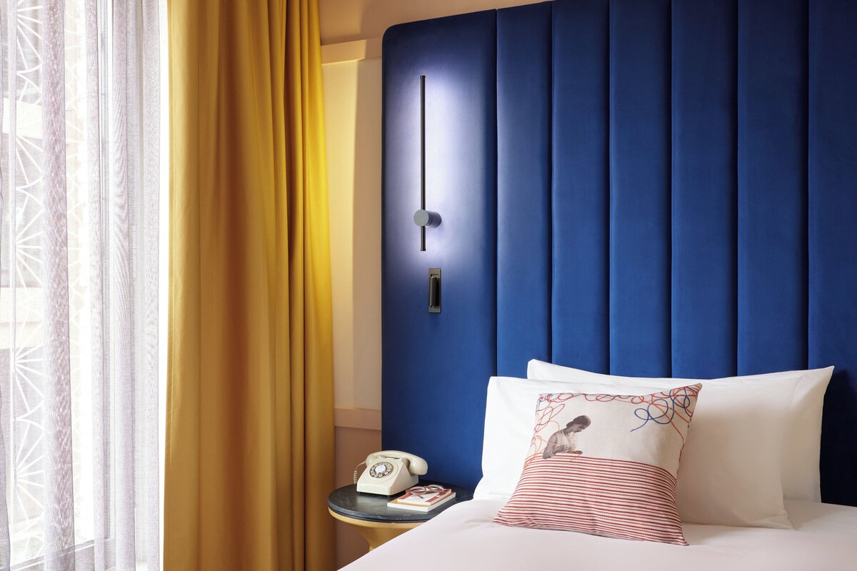A hotel room with a blue headboard, a bedside telephone, a small table with a book, and a window with curtains.