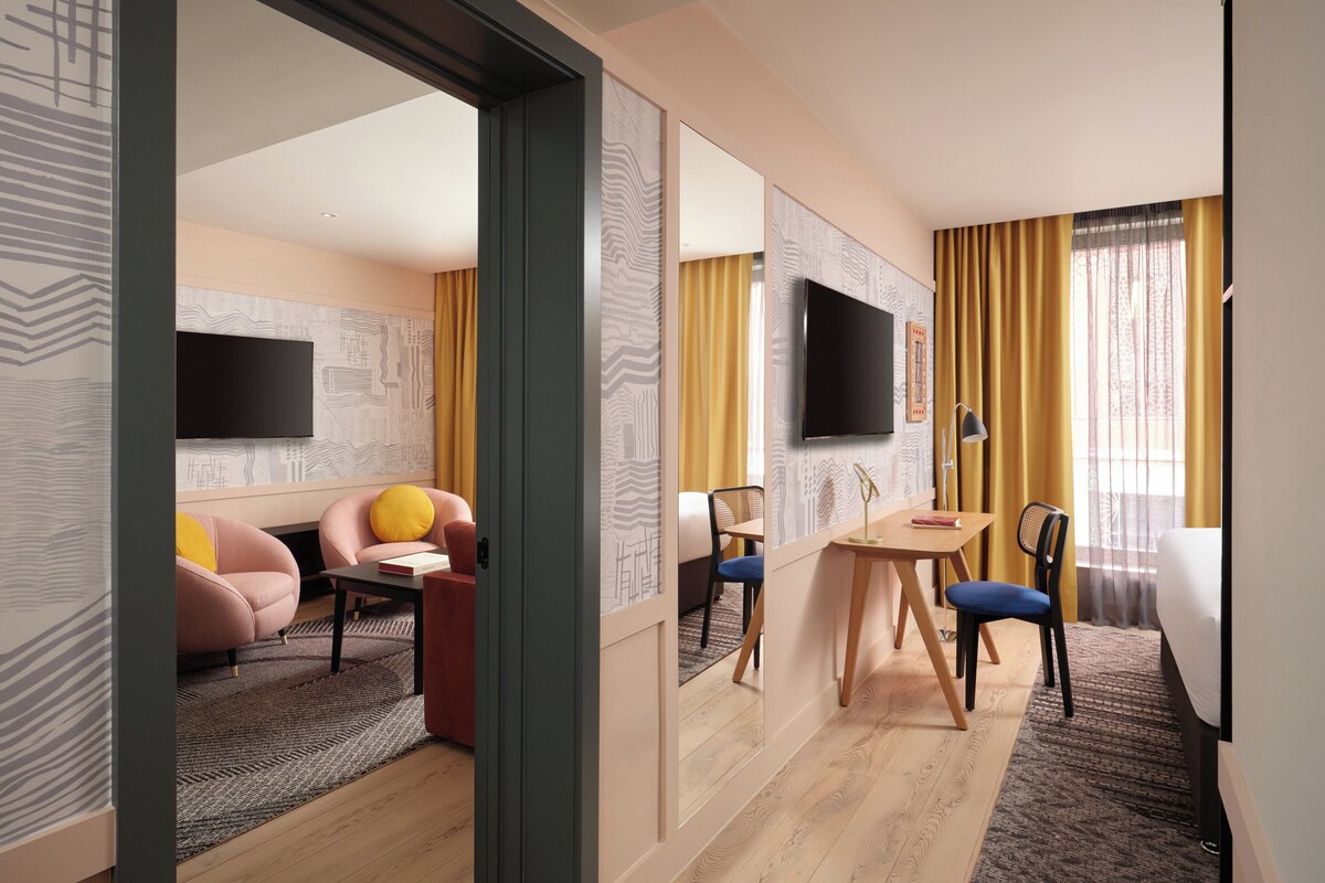 A hotel room with a flat-screen TV, a bed, a desk with a chair, a sofa, and a dining table.
