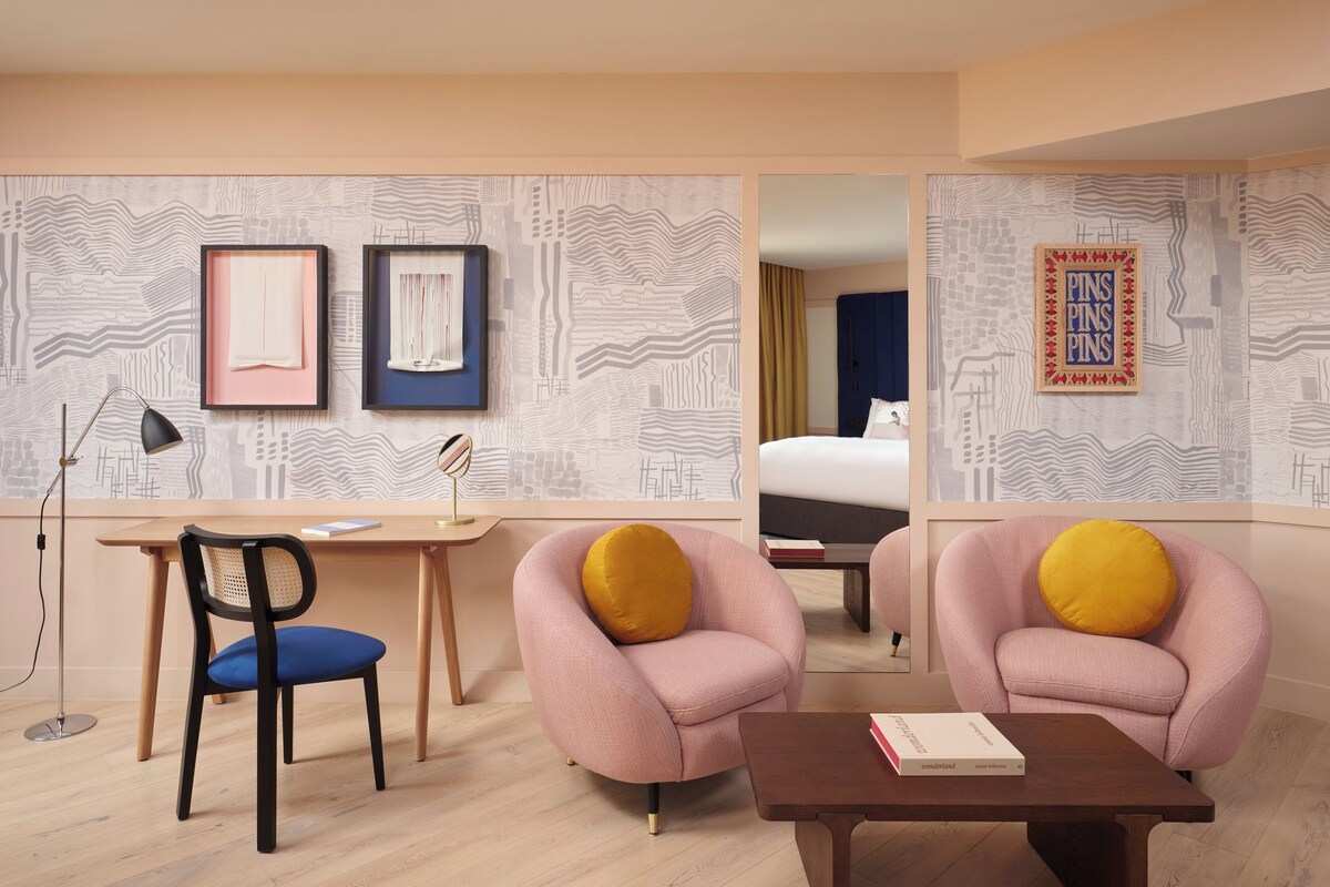 Una habitación de hotel moderna con un escritorio de madera, dos sillones rosados con cojines amarillos, una silla azul y una mesita.