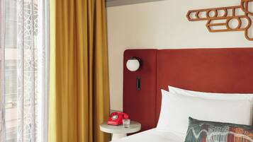 A hotel room with a bed, a bedside table with a red rotary phone, a glass of water, and a decorative wall piece.