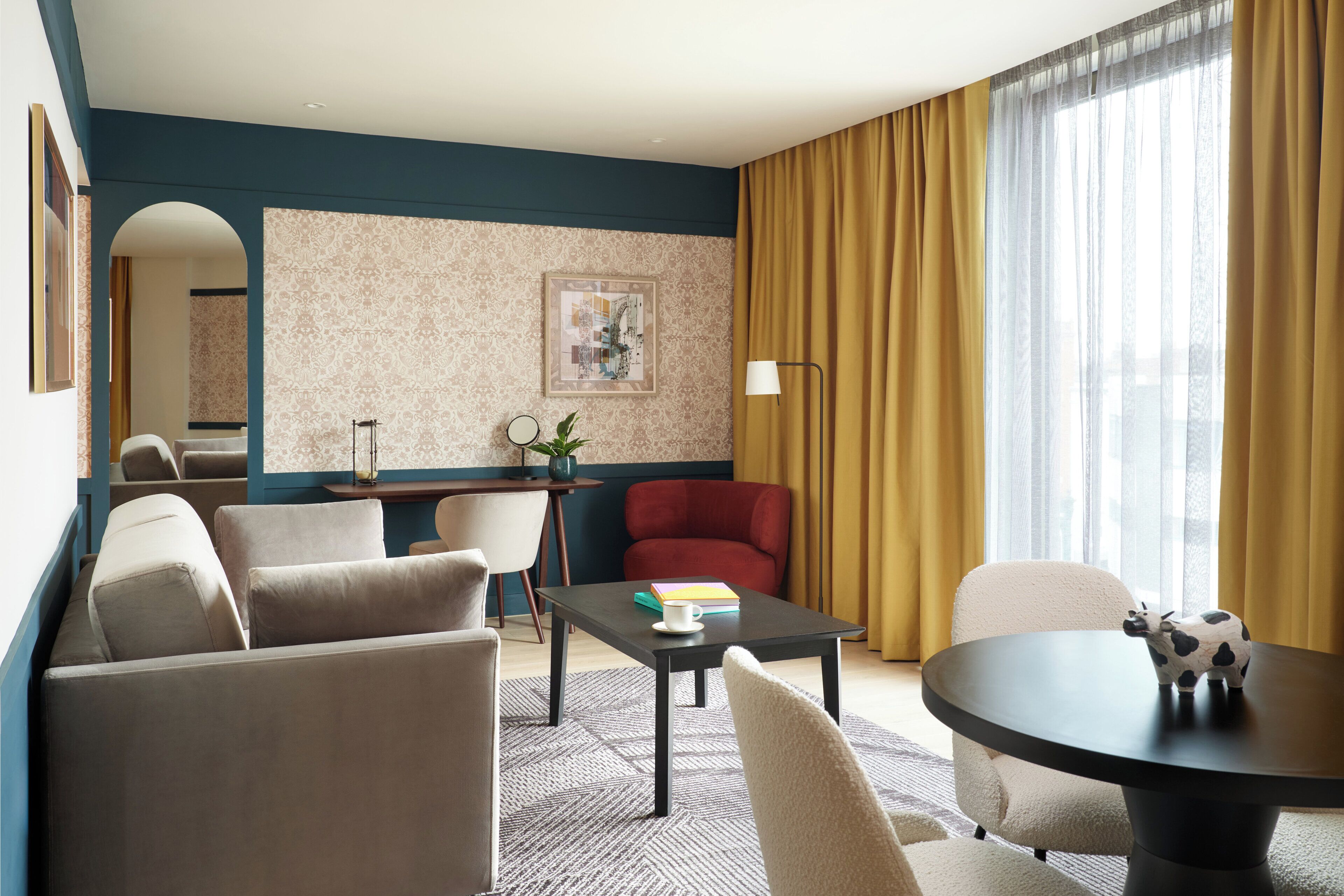 Quarto de hotel moderno com sofá, mesa de jantar redonda, poltrona vermelha, pequena escrivaninha com luminária e uma janela grande com cortinas.