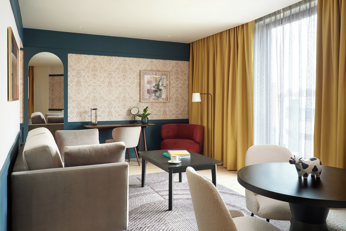 Una habitación de hotel moderna con un conjunto de sofá, una mesa de comedor redonda, un sillón rojo, un escritorio pequeño con una lámpara y un ventanal con cortinas.