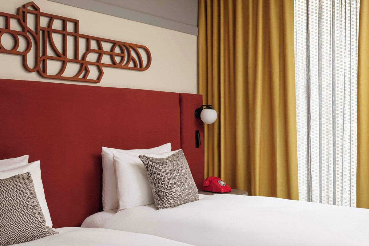 Habitación de hotel con cabecera roja, dos camas blancas, almohadas estampadas, teléfono rojo e lámpara fijada a la pared.