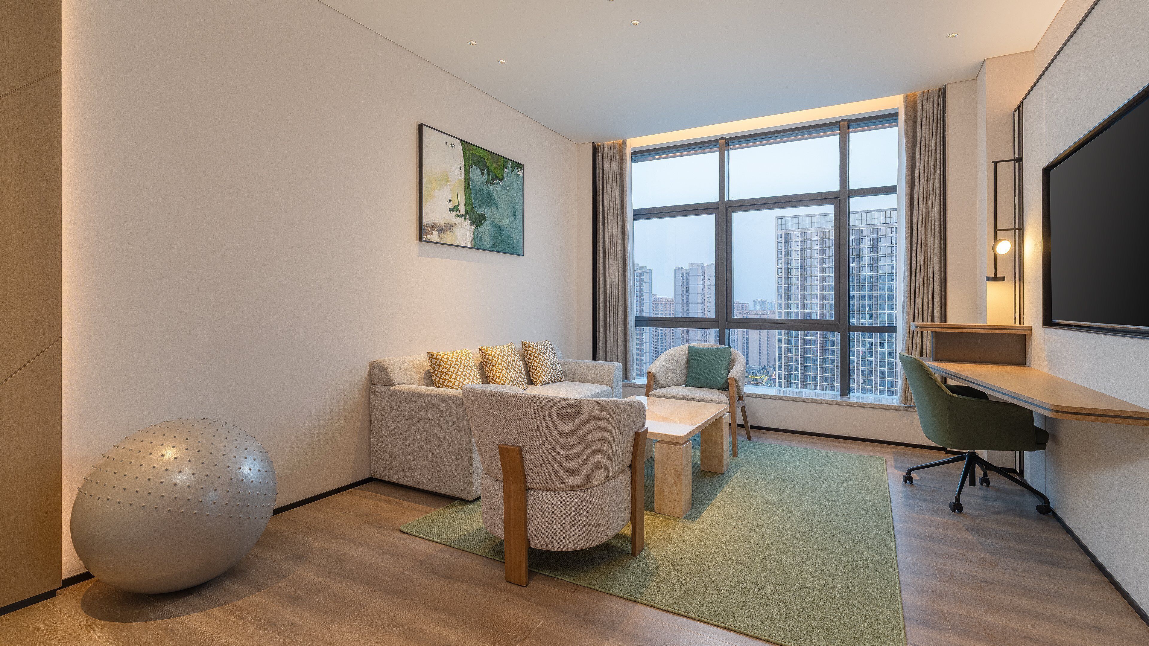 A modern hotel room with a large window, a sofa, a desk, and a green exercise ball.