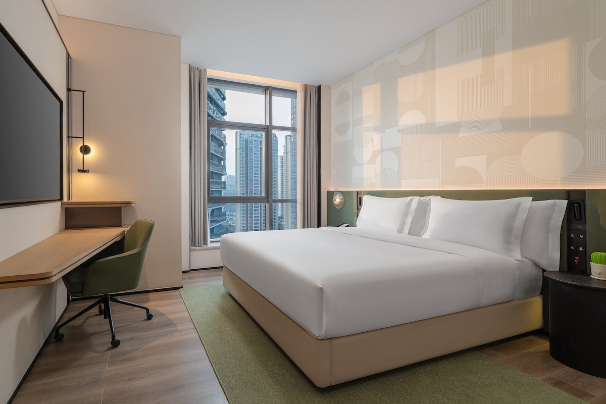 A hotel room with a bed, desk, chair, and a large window with a city view.