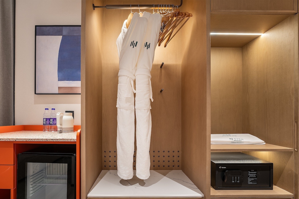A hotel room with a bed, a small table with a water bottle and a coffee maker, a robe hanging on a hook, and a minibar.