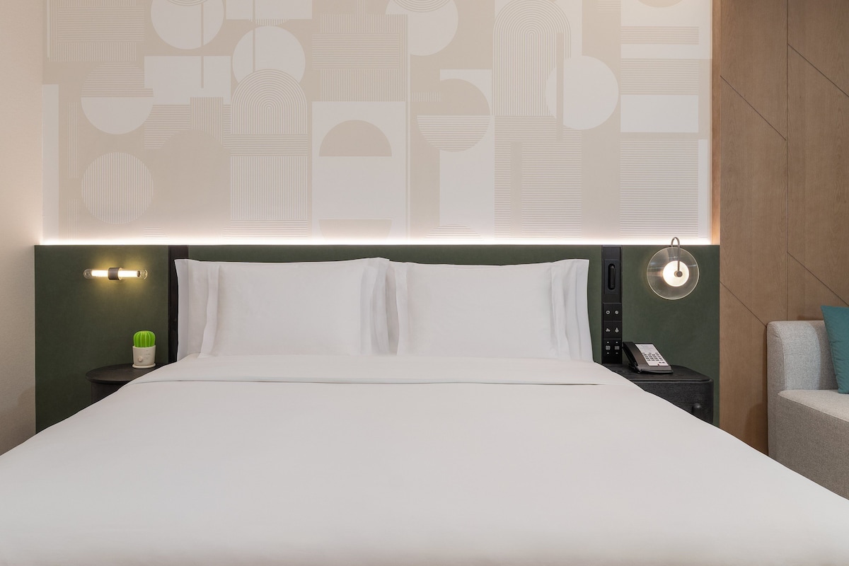 A neatly made bed with white linens, a bedside lamp, a small green plant, and a wall-mounted phone on a nightstand.
