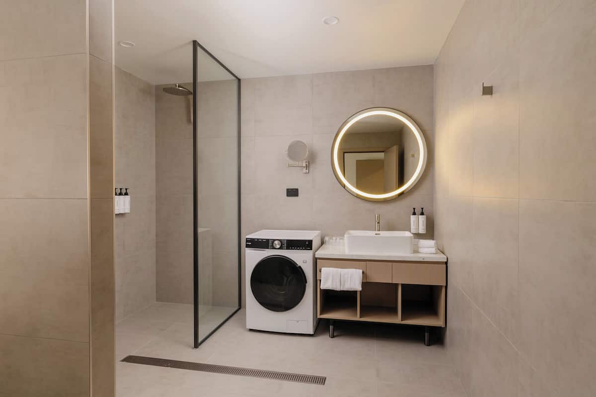 A modern bathroom with a glass shower enclosure, a built-in washing machine, a sink with a mirror, and a towel rack.
