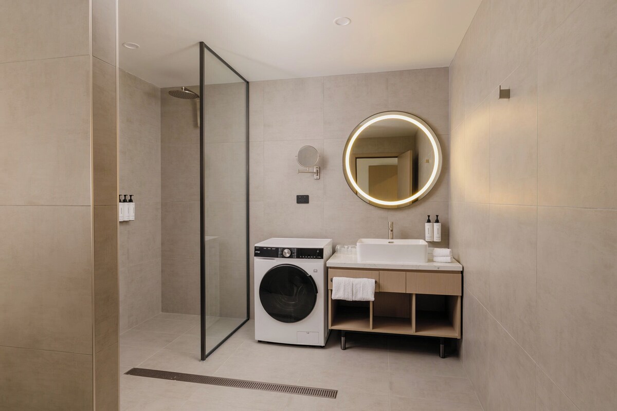 A modern bathroom with a glass shower enclosure, a built-in washing machine, a sink with a mirror, and a towel rack.