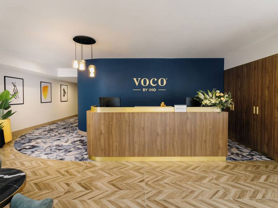 A reception desk with a wooden finish and a blue wall behind it, featuring the logo 'VOCO BY IHG'.