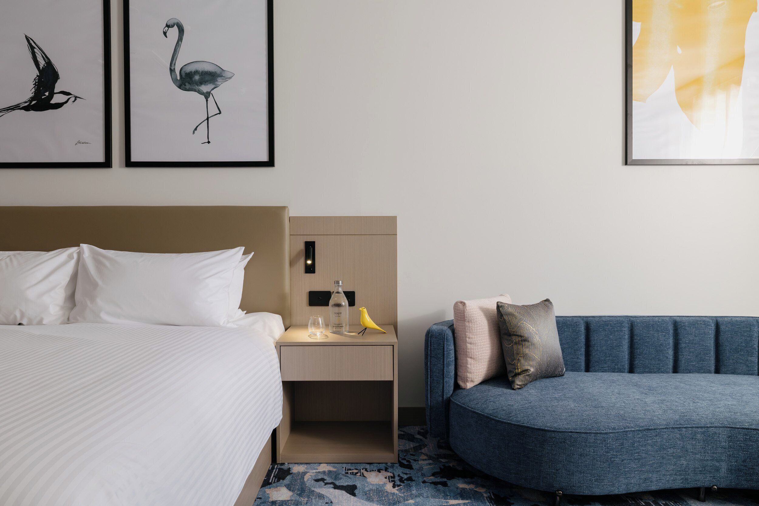 A hotel room with a bed, a nightstand, a blue sofa, and two framed wall art pieces.