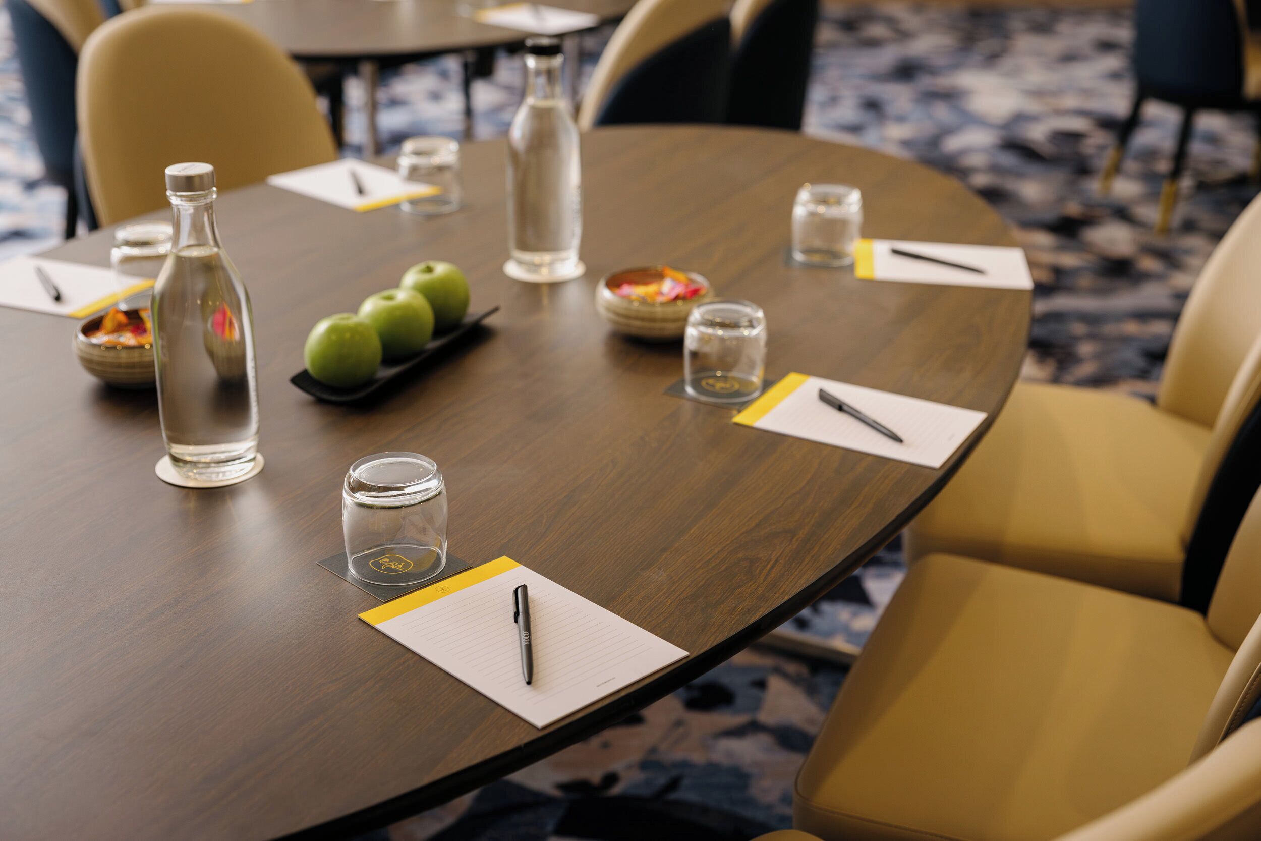 Ein Konferenztisch mit einer Flasche, Gläsern, Äpfeln und einem Stift auf einem Notizblock.