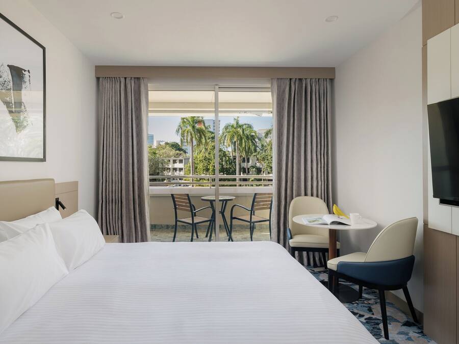 Habitación de hotel con cama, televisión, balcón con sillas y mesa, y vistas a palmeras y edificios.