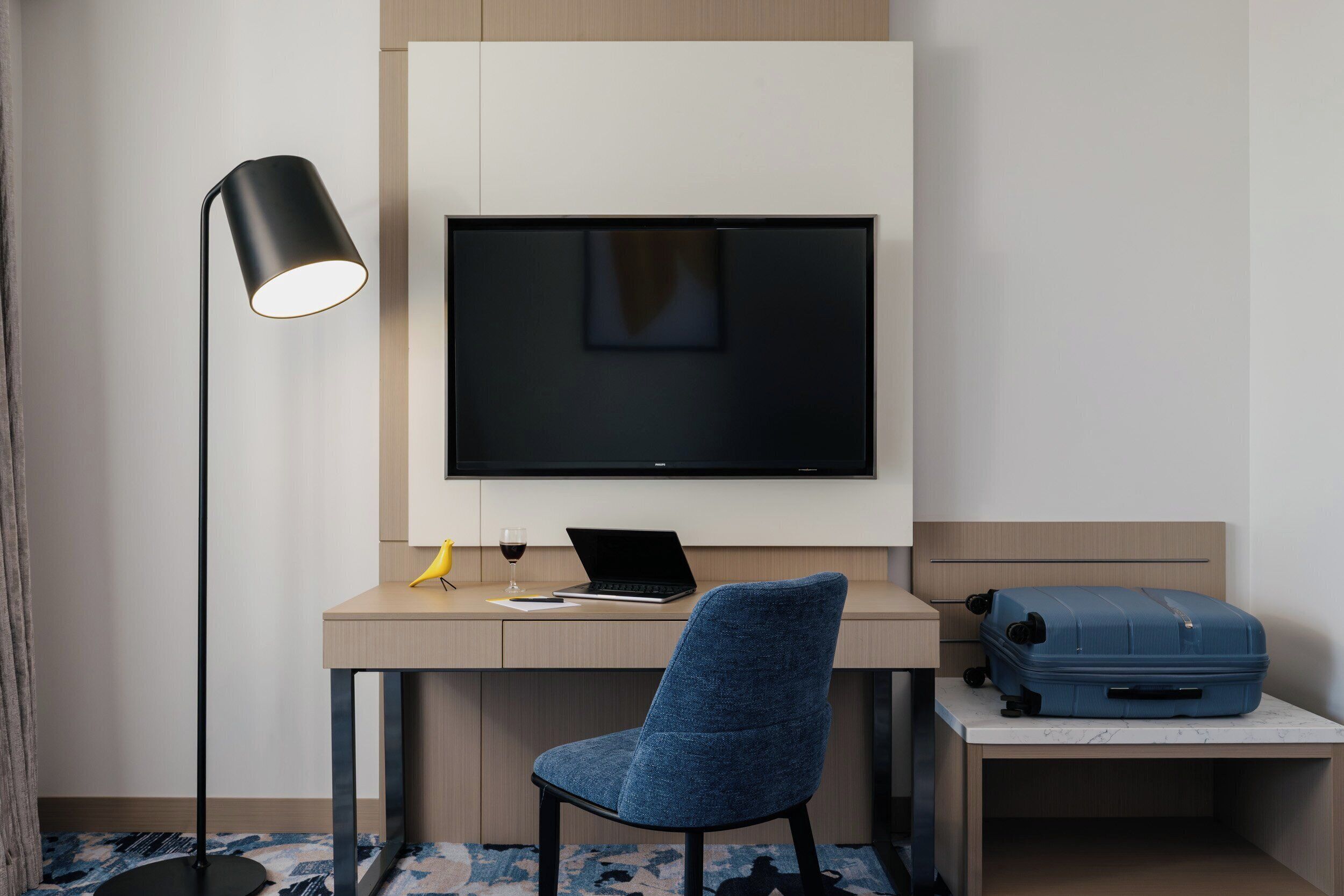 Een hotelkamer met een bureau, een stoel, een televisie, een vloerlamp en een koffer.