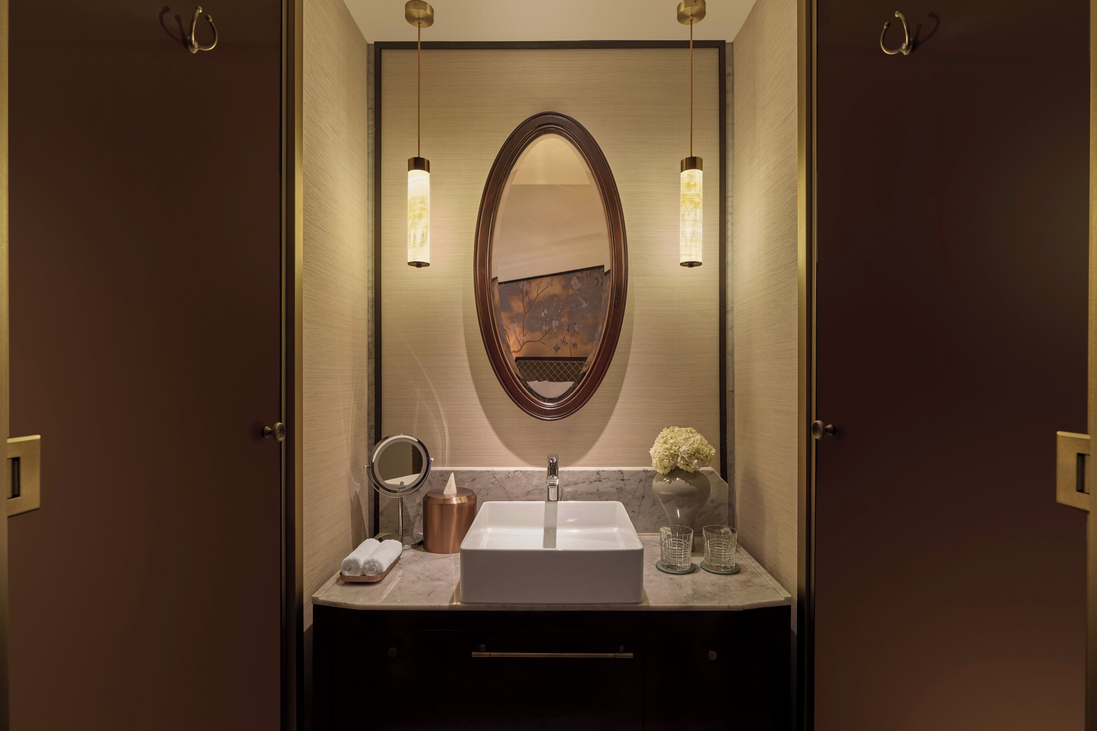 Une salle de bain avec un grand miroir ovale, un lavabo blanc, un distributeur de serviettes et deux lampes fixées au mur.