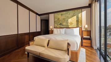 A bedroom with a large bed, a sofa, a nightstand, a lamp, and a city view through large windows.