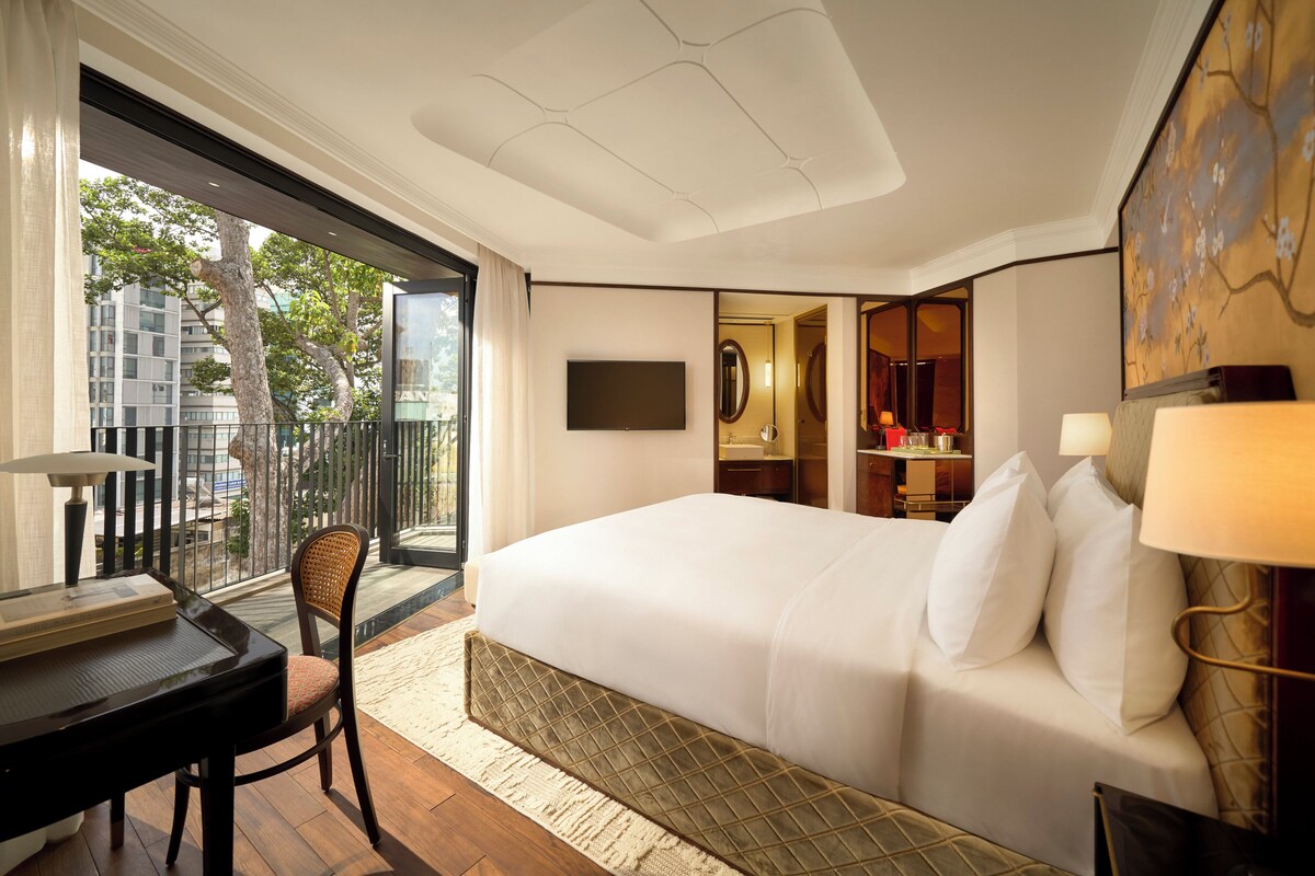 A modern hotel room with a large bed, a desk with a lamp, a chair, and a view of the city through a large window.